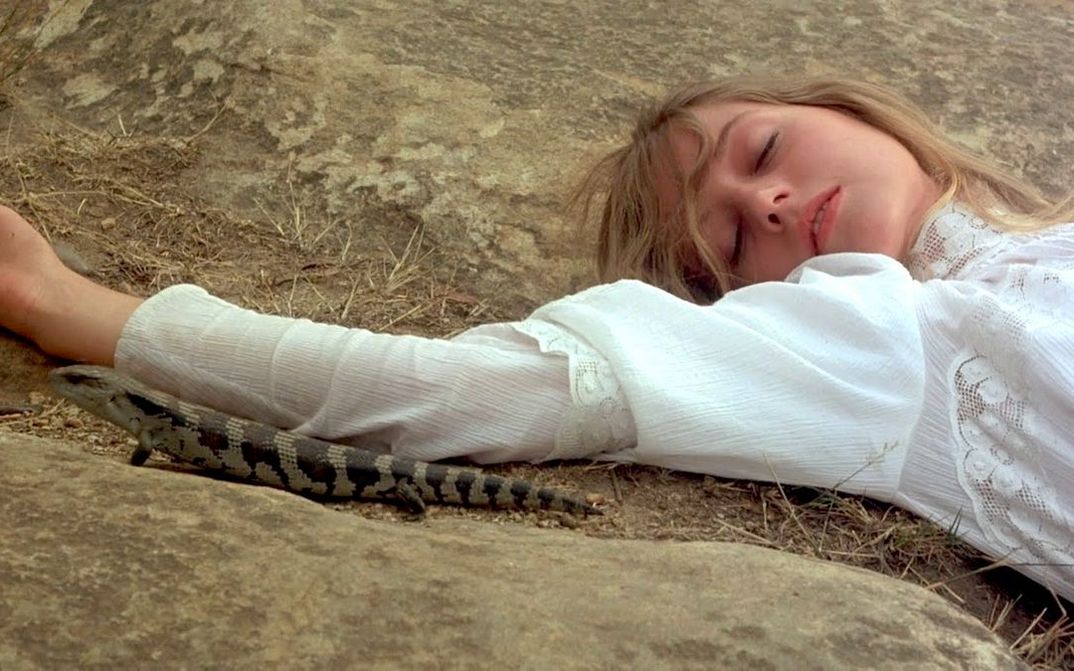 Film still from PICNIC AT HANGING ROCK: A woman lies on a rocky floor with her eyes closed. She is wearing a white dress. A lizard sits next to her arm.