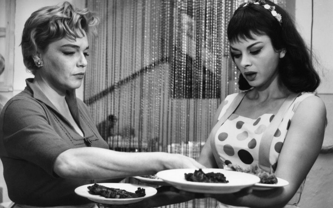 Film still from ADUA E LE COMPAGNE: Two women are standing in a restaurant, one of them is carrying four plates.