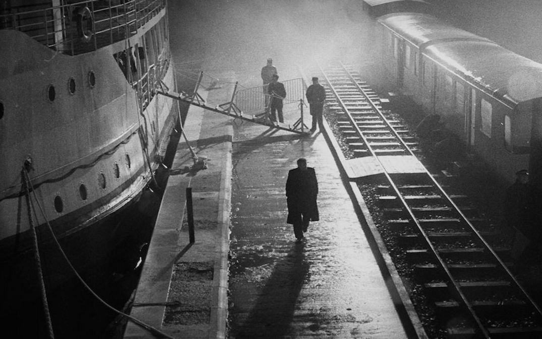 Film still from MAN FROM LONDON: Night-time shot with strong light contrasts. A few people can be seen between a landing stage and the railroad tracks.