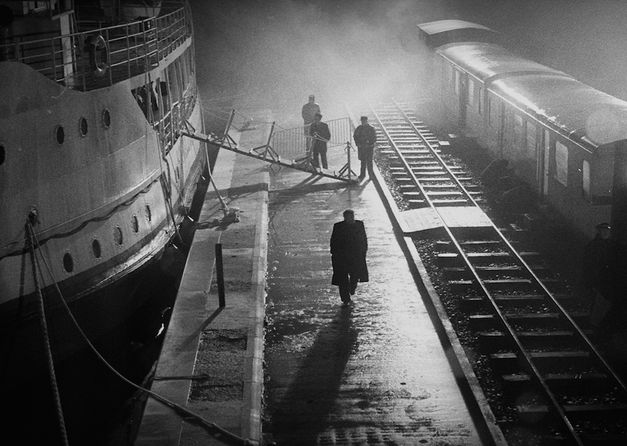 Film still from MAN FROM LONDON: Night-time shot with strong light contrasts. A few people can be seen between a landing stage and the railroad tracks.