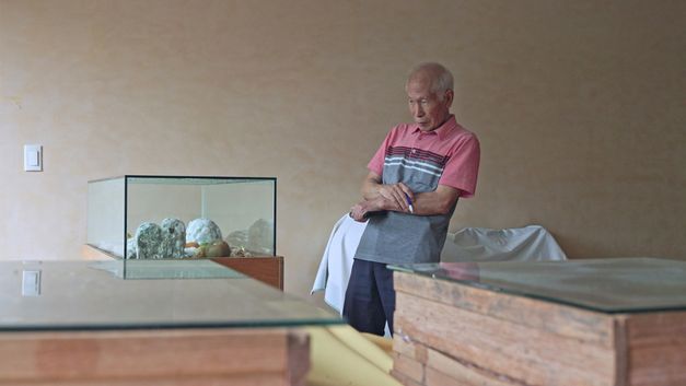 Film still from Jeamin Cha’s film “Photosynthesizing Dead in Warehouse”. A middle-aged man observes the decomposition of organic elements placed under a glass display case.