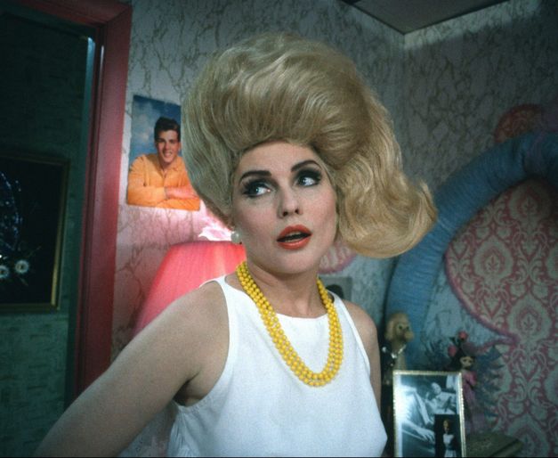 Filmstill from HAIRSPRAY: A woman with tousled blonde hair and a yellow pearl necklace looks to the left and stands in a room with eye-catching wallpaper and posters in the background.