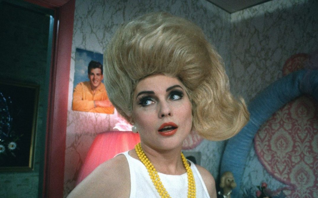 Filmstill from HAIRSPRAY: A woman with tousled blonde hair and a yellow pearl necklace looks to the left and stands in a room with eye-catching wallpaper and posters in the background.