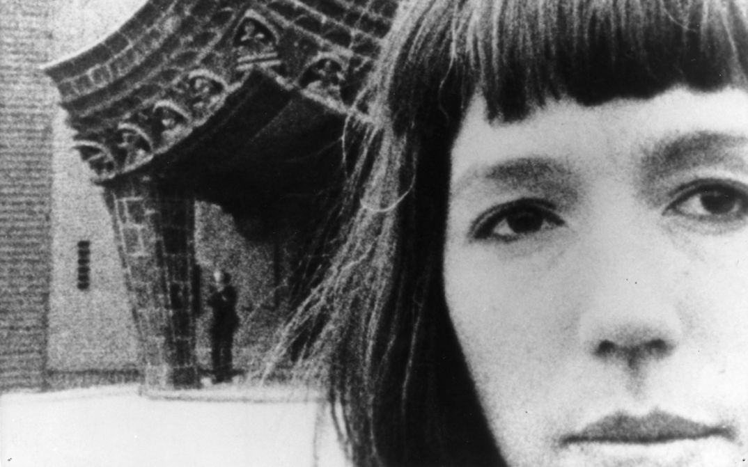 Film still from JOURNEYS FROM BERLIN/1971: We see the large face of a woman, in the background is the entrance to a building.