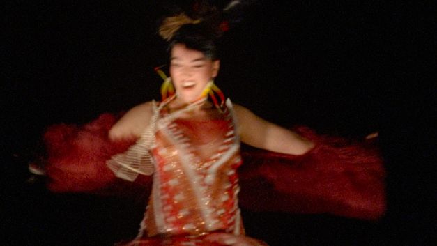 Film still from Caroline Monnet’s film “Pidikwe”. A person appears to be dancing or spinning, with arms spread wide and a radiant expression. The blurred image captures the rapid movement of the moment. They are wearing a contemporary creation that dialogues with Native American clothing heritage, blending red and golden tones, with geometric patterns and fringes that wave with the movement.