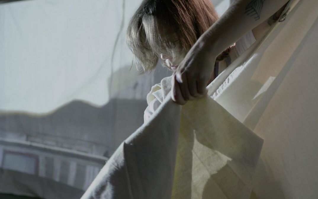 Filmstill from the film "The Song of the Shirt" by Kerstin Schroedinger. You see a person stretching out a white fabric.
