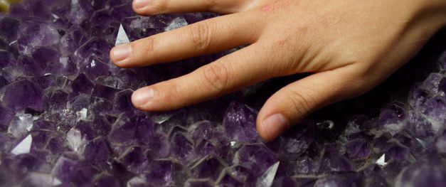Hand berührt violette Kristalle mit scharfkantiger, unregelmäßiger Oberfläche.