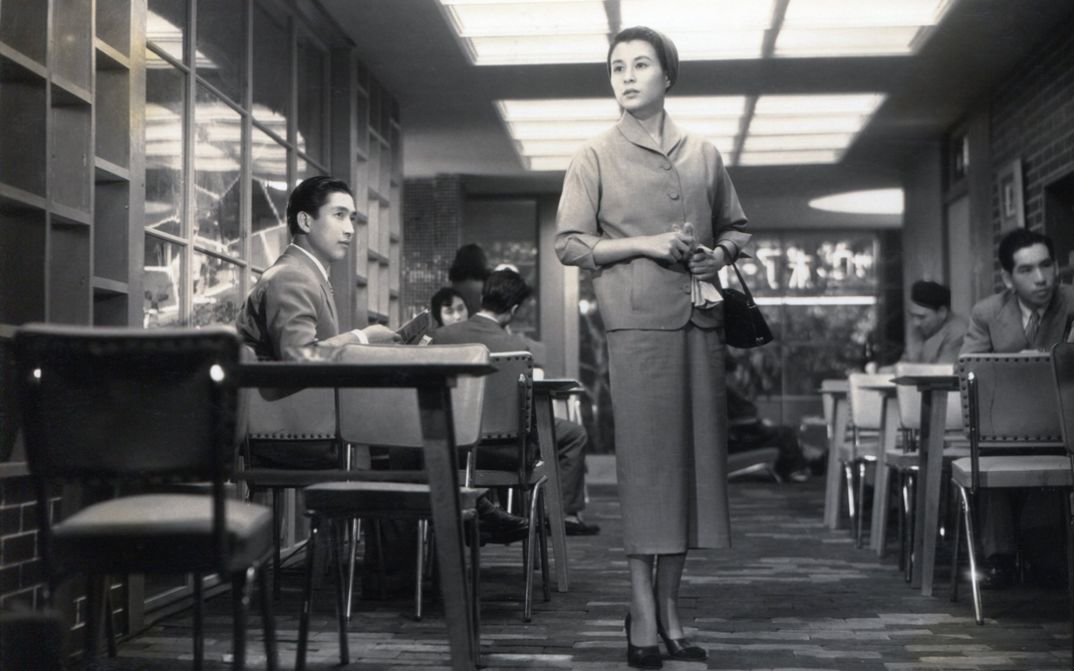 Film still from KOIBUMI: A woman stands in a pub and looks around searchingly.