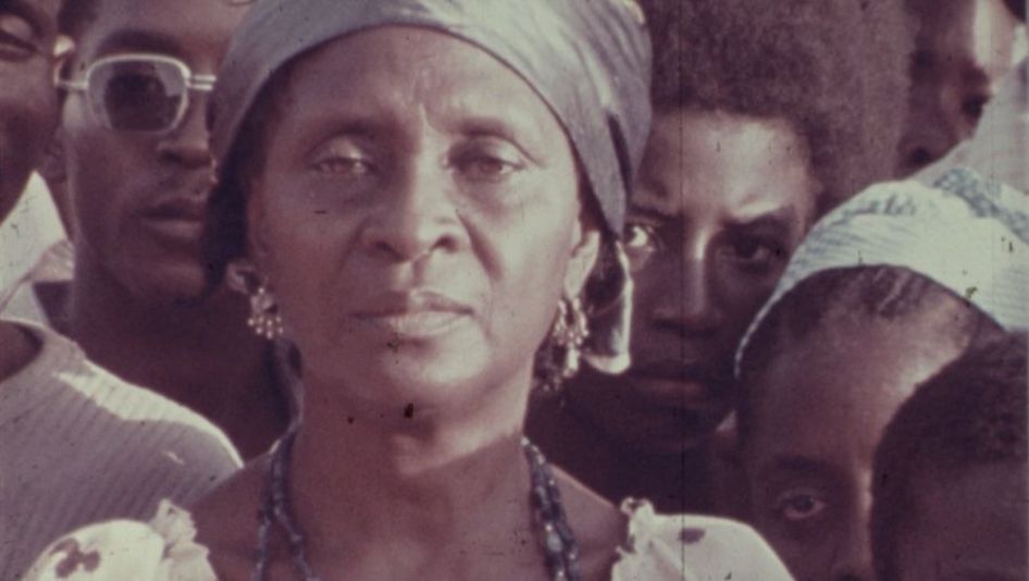 Filmstill aus The Return of Amílcar Cabral: Eine Frau blickt mit ernstem Gesicht in die Kamera. Um sie herum stehen weitere Menschen.