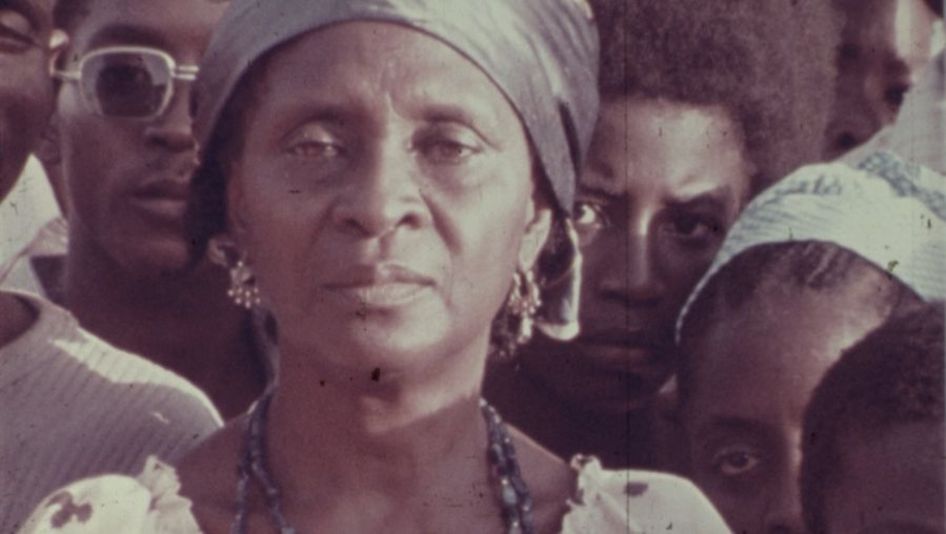 Film still from The Return of Amílcar Cabral: A woman looks into the camera with a serious face. Other people are standing around her.