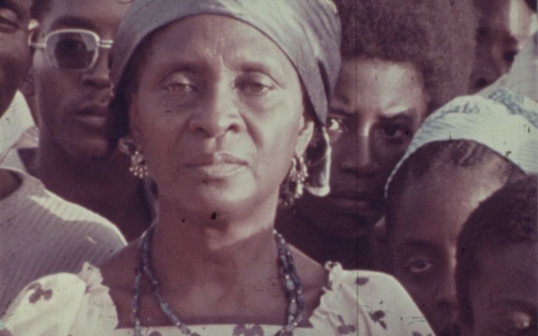 Film still from The Return of Amílcar Cabral: A woman looks into the camera with a serious face. Other people are standing around her.