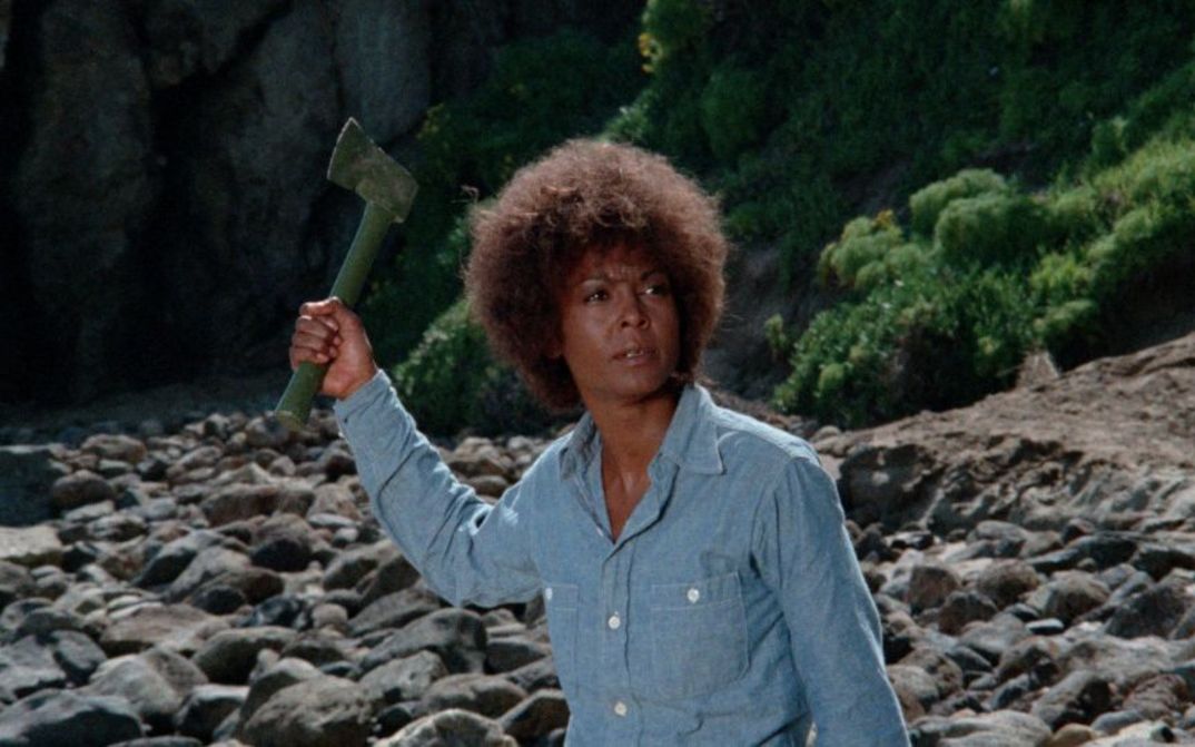 Film still from TERMINAL ISLAND: A woman holds an axe in her raised hand and stands in a barren landscape.