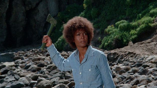 Film still from TERMINAL ISLAND: A woman holds an axe in her raised hand and stands in a barren landscape.