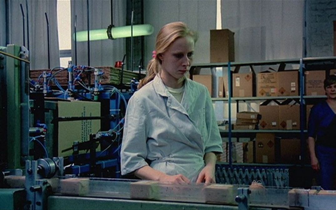Film still from THE MATCH FACTORY GIRL: A young woman stands at an assembly line in a factory.