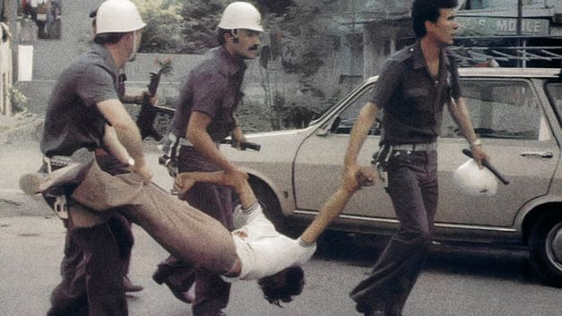 Three uniformed men (policemen) carry a man by his arms and legs.  