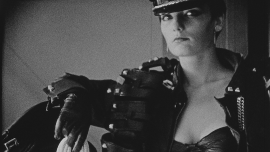 Film still from MANO DESTRA: A woman dressed in leather and holding a rope looks into the camera.