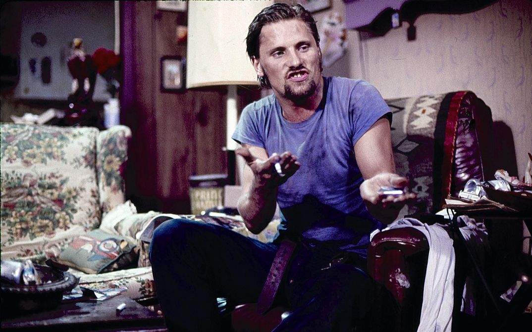 Film still from SALVATION! A man in a blue shirt and jeans sits on an armchair and gesticulates strongly with his hands. He speaks with a roar. A living room in the background.