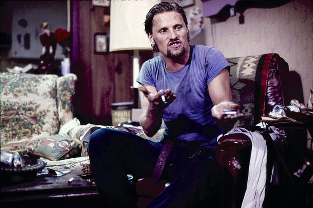 Film still from SALVATION! A man in a blue shirt and jeans sits on an armchair and gesticulates strongly with his hands. He speaks with a roar. A living room in the background.