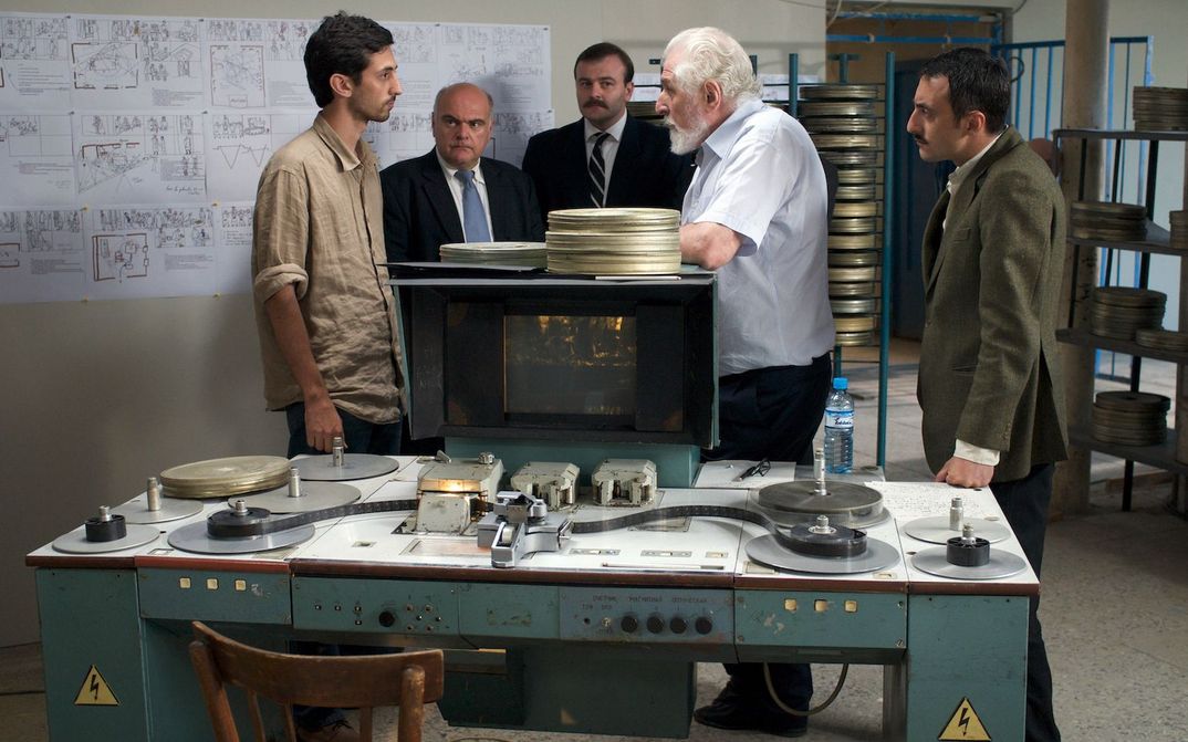 Film still from CHANTRAPAS: A group of men discussing in a room. An editing table with a 35mm film can be seen in the foreground. The room is full of film cans and a drawn storyboard is hanging.
