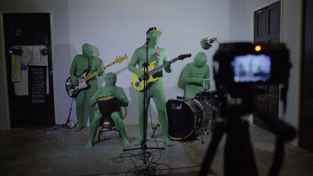 A rock band in green full-body suits, which also cover their heads, stand in formation in front of a digital camera. They hold their instruments in their hands. 