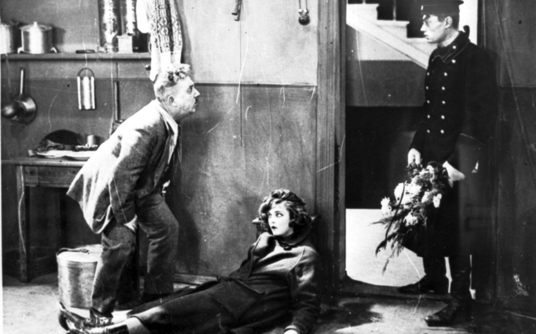 Film still from DAS MÄDCHEN MIT DER HUTSCHACHTEL: A young woman is half lying and half sitting, leaning against the wall, in an apartment. To her left is an older man looking at a younger man in uniform standing in the doorway with a bouquet of flowers in his hand.