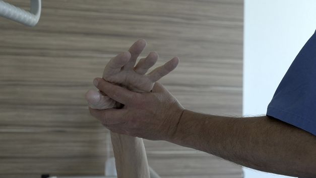 Ein Arm einer Krankenpfleger*in hält die Hand eine*r Patient*in hoch vor einer Wand mit Holzfournier.