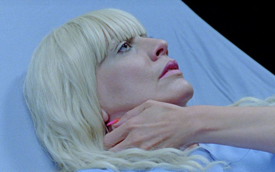 Film still from SIRENS CALL by Miri Ian Gossing and Lina Sieckmann. The image shows a close-up of a blonde woman in a hospital bed. She is clutching her neck with her hand.