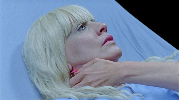 Film still from SIRENS CALL by Miri Ian Gossing and Lina Sieckmann. The image shows a close-up of a blonde woman in a hospital bed. She is clutching her neck with her hand. 