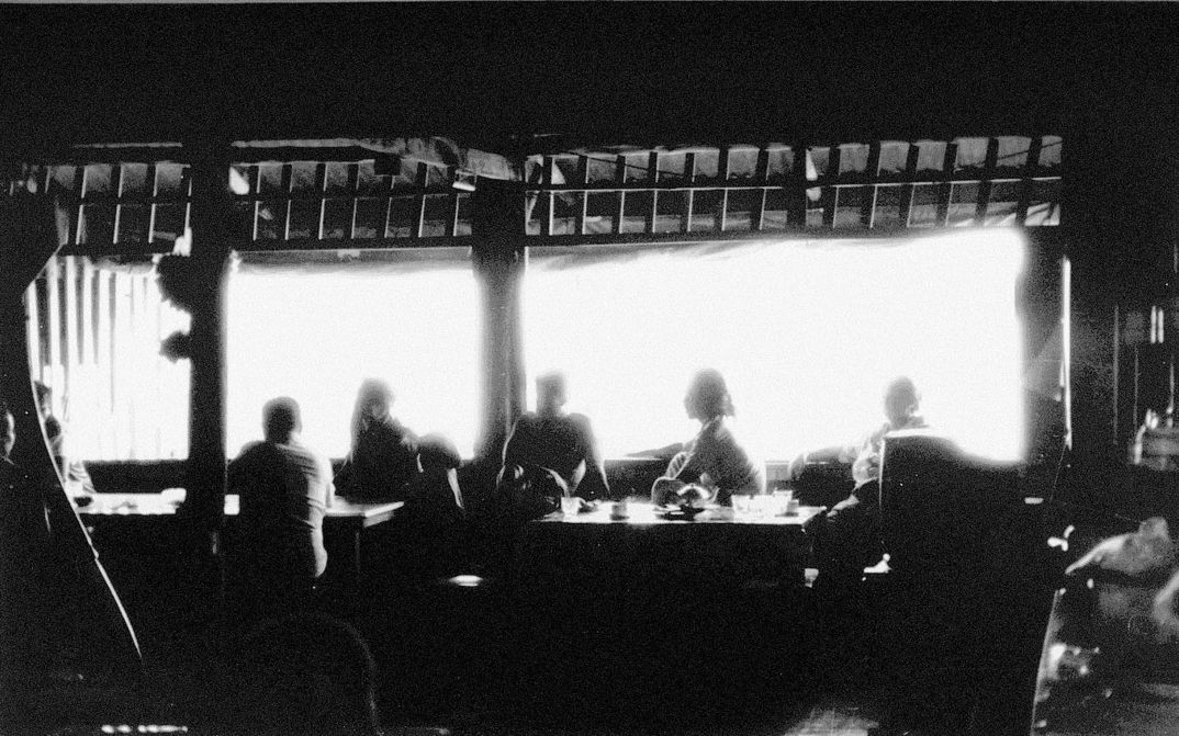 Filmstill aus MYSTERIOUS OBJECT AT NOON: Eine leicht verschwommene Aufnahme einer Gruppe von Menschen, die an einem langen Tisch sitzen. Durch ein Fenster strömt helles Licht in den Raum.