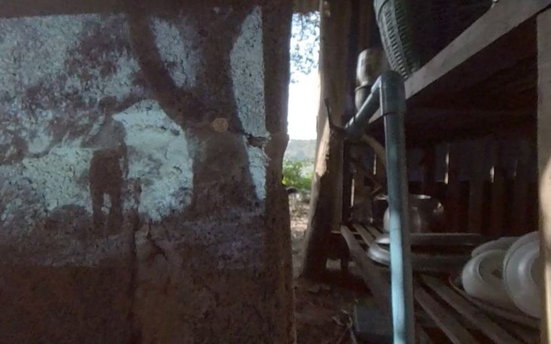 Filmstill aus dem Film "Trip After" von Ukrit Sa-nguanhai. Der Ausschnitt einer Kochstelle, ein Wasserhahn und - auf eine Holzwand projiziert - das Bild einer älteren Person unter einem Baum.
