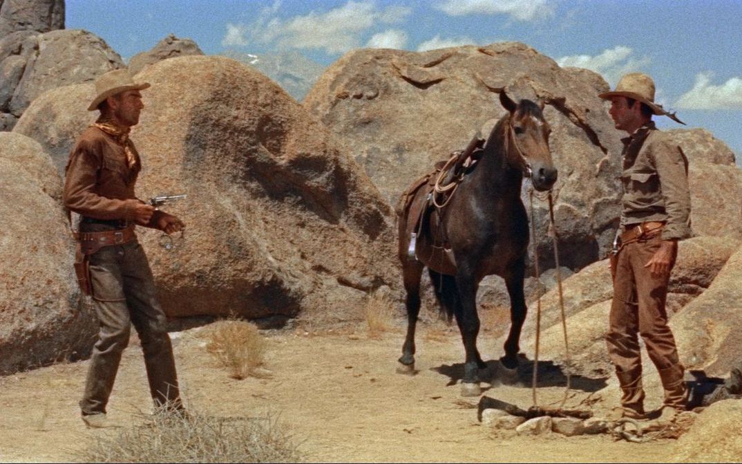 Film still from RIDE LONESOME: Two cowboys face each other in a rocky desert landscape, with a horse standing between them. One is pointing his revolver at the other.