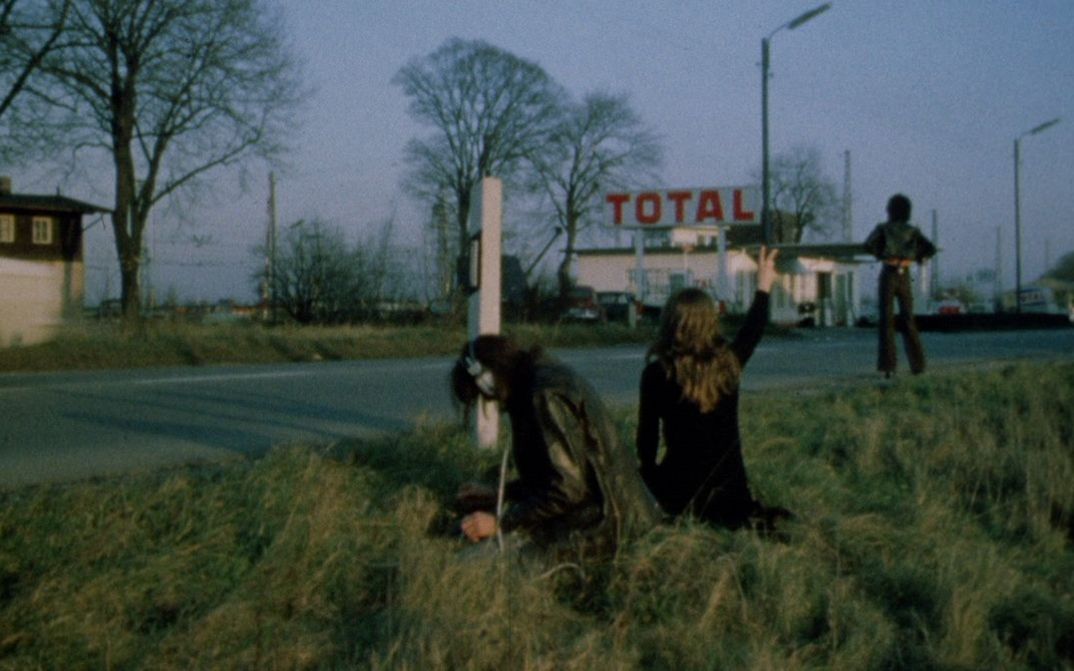 Film still from LEAVE ME ALONE. Three people are standing on the side of a road, with a gas station in the background.