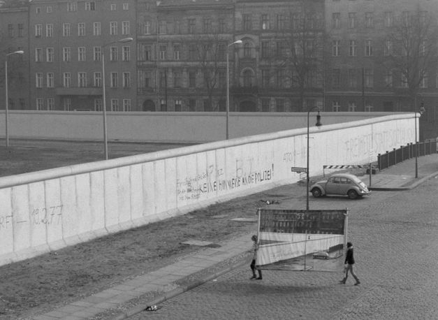 Film still from REDUPERS: View of the Berlin Wall in Kreuzberg. Small, you can see two women carrying a photo wall with this very view.