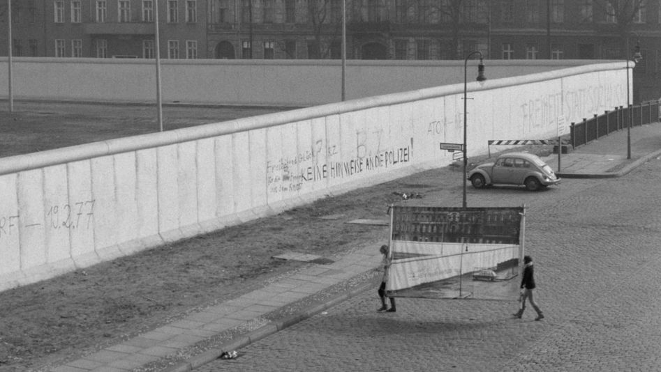 Film still from REDUPERS: View of the Berlin Wall in Kreuzberg. Small, you can see two women carrying a photo wall with this very view.