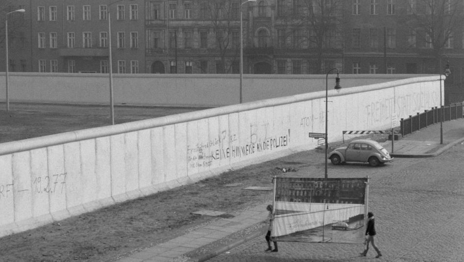 Filmstill aus REDUPERS: Ansicht der Berliner Mauer in Kreuzberg. Klein sieht man zwei Frauen, die eine Fotowand mit ebendieser Ansicht herantragen.