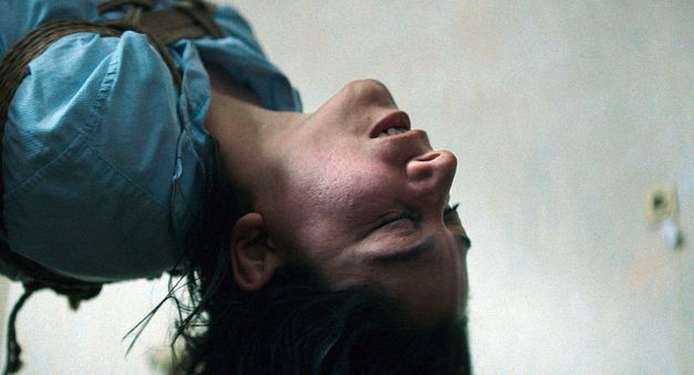 Nahaufnahme einer Person mit zurückgeneigtem Kopf, die mit Seilen oder Gurten fixiert zu sein scheint, vor hellem Hintergrund.