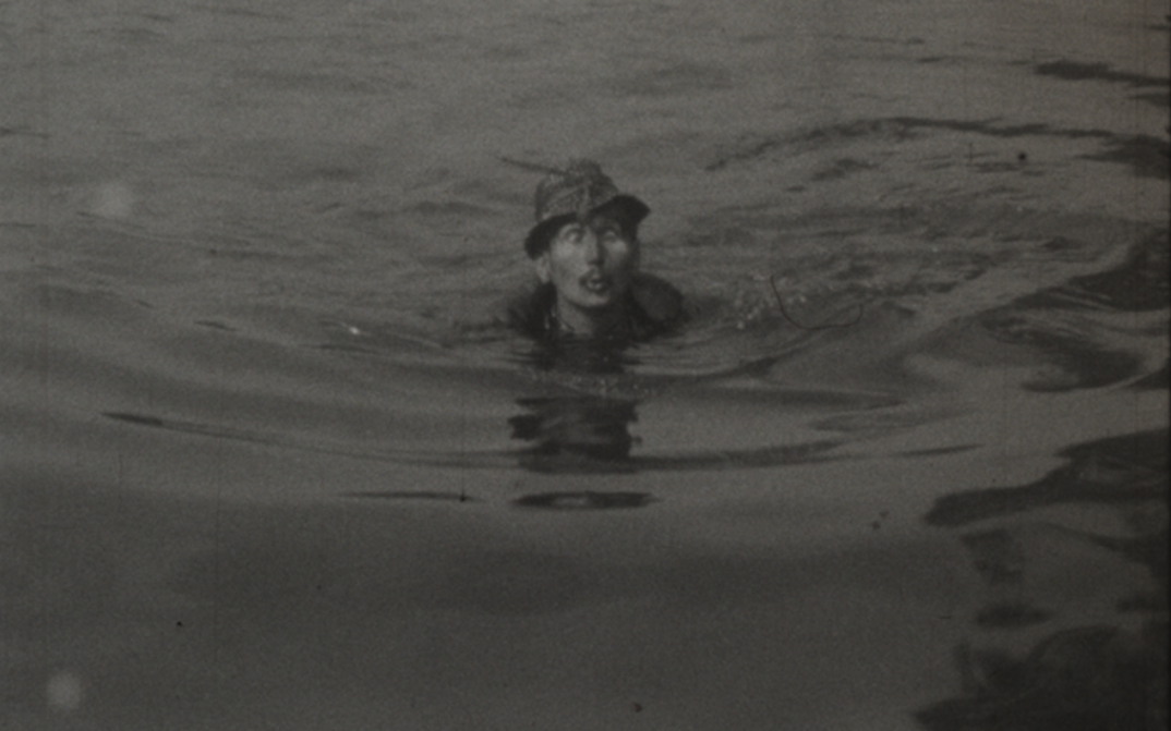 Film still from HASTRMAN: Ein Mann mit Hut schwimmt in in einem Gewässer.