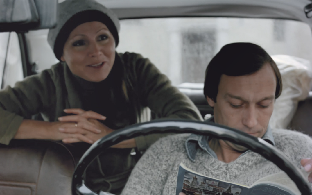 Film still from FLIGHTS IN DREAM AND REALITY: A man and a woman in a car, he sits in the front reading a magazine, she sits in the back leaning forward.