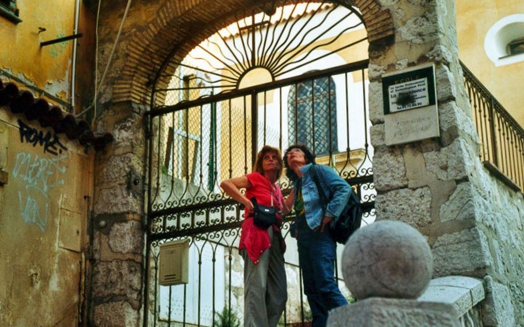 Film still from MIMI: Two women are standing on the street in front of an archway with a fence.