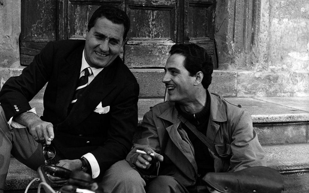 Film still from LO SCAPOLO: Two men are sitting on a staircase laughing and smoking.