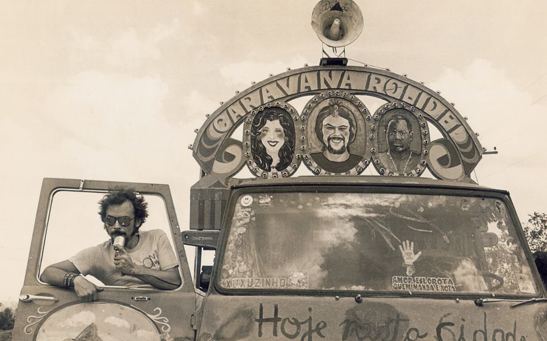 Film still from BYE BYE BRASIL: A man stands in the doorway of a painted car equipped with a loudspeaker.