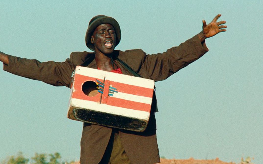 Film still from LE FRANC: A man stands in the landscape with his arms outstretched and a painted wooden box hanging around his neck.