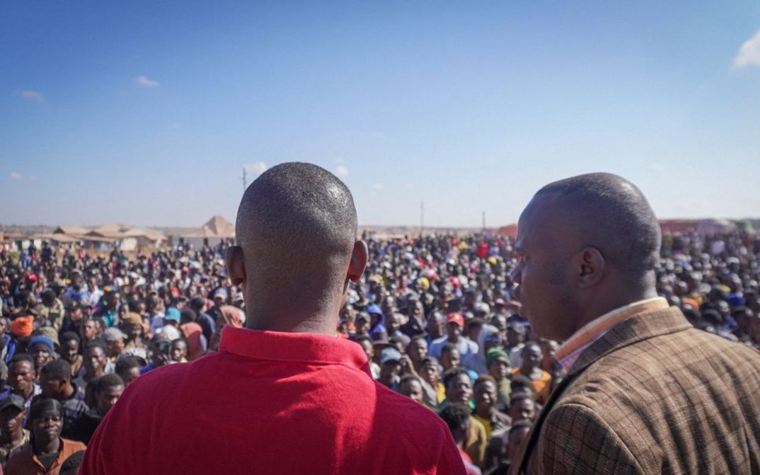 Film still from Petna Ndaliko Katondolo’s film “Mikuba”. Two men, seen from behind, overlook a crowd gathered below.