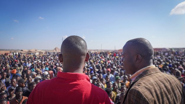 Film still from Petna Ndaliko Katondolo’s film “Mikuba”. Two men, seen from behind, overlook a crowd gathered below.