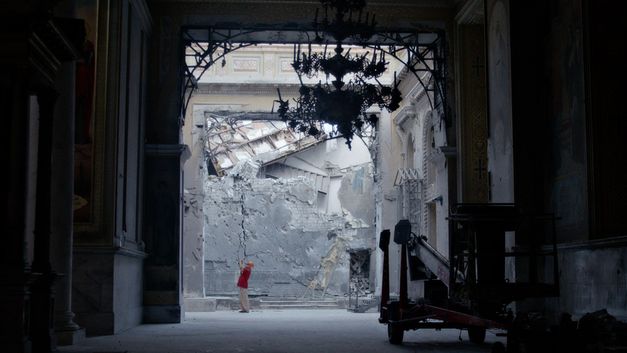 We are standing in a passageway and can see a damaged wall in the background. A man is standing at the back taking photographs.