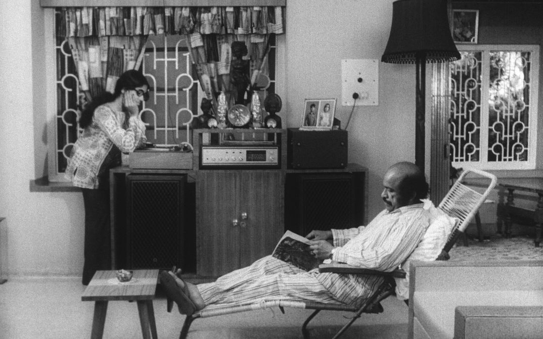 Film still from THAMP. A man and a woman in a living room, he is sitting in a deck chair reading, she is standing at the record player listening to the music.