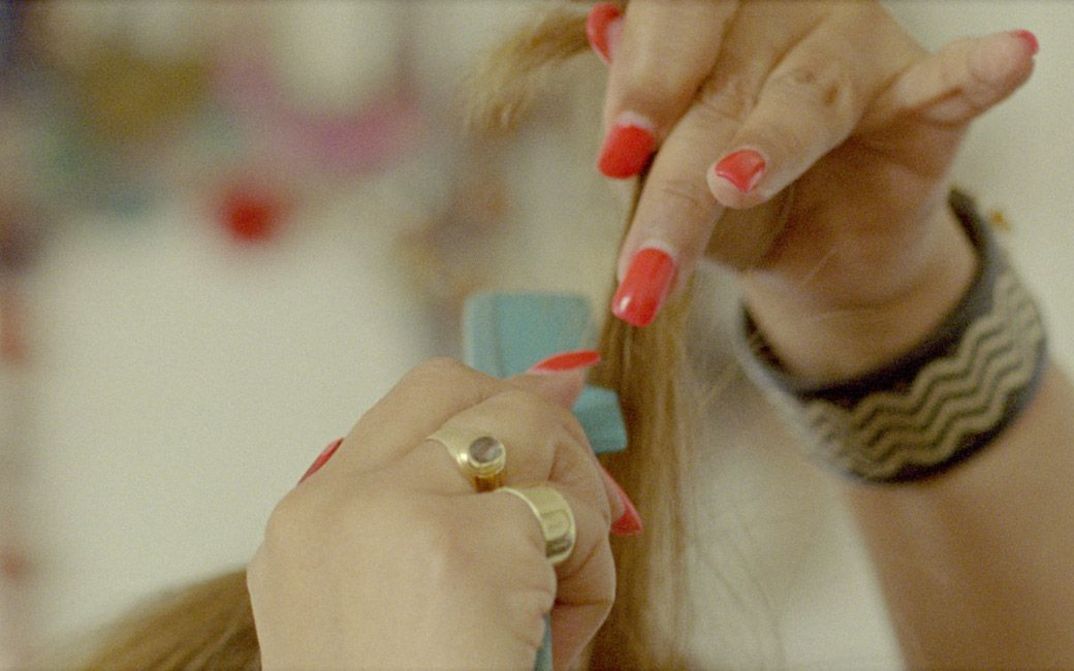Filmstill aus „My Two Voices“ von Lina Rodriguez. Nahaufnahme von zwei Händen mit rot lackierten Fingernägeln. Die eine Hand hält blondes Haar in die Höhe, die andere Hand fährt mit einem blauen Kamm durch das Haar.