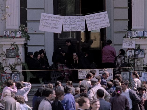 A protest scene: a crowd in front of a building, with placards and photos of faces. 