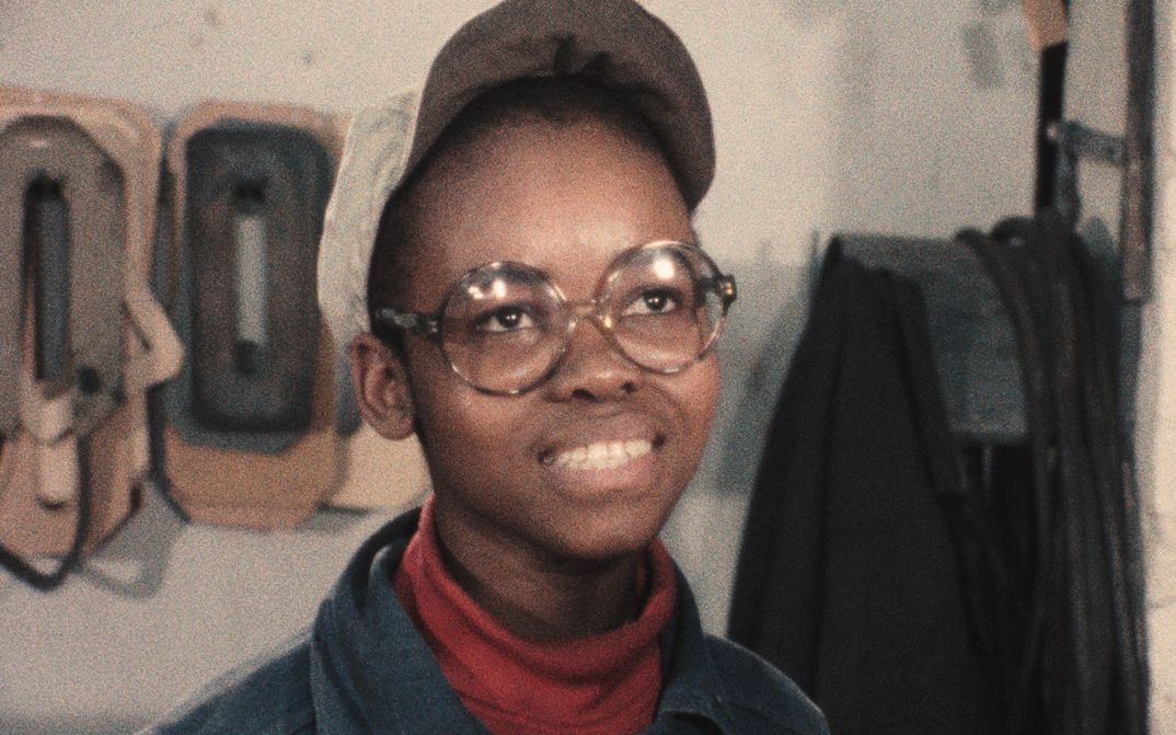 Film still from GLÜCK AUF – BILDER EINER BEGEGNUNG: A young person with a white cap and round glasses is grinning at the camera.