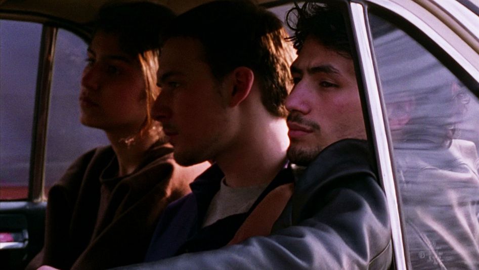 Film still from GESCHWISTER: Three young people sit in the back seat of a car with the window open.